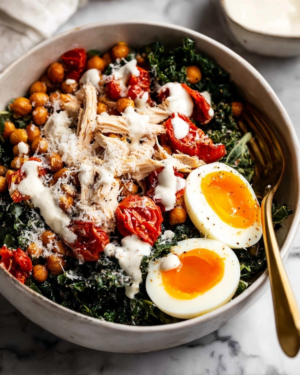 A bowl with four main layers is shown on a white marbled texture. The bottom layer is dark green kale with a rough, leafy texture that fills most of the bowl. On top of the kale, there are pieces of shredded white chicken and bright red roasted cherry tomatoes spread unevenly. On one side of the bowl, there are two halves of a soft-boiled egg with a bright orange yolk and white edges. The top layer is golden brown roasted chickpeas scattered in the center, covered partially with a smooth, creamy white dressing. The dish is sprinkled with finely grated white cheese, adding a light texture over the chickpeas and chicken. A gold spoon rests inside the bowl near the chicken. photo taken with an iphone --ar 4:5 --v 7