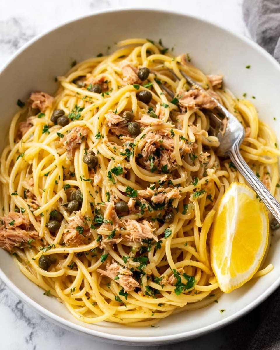 A white bowl filled with a tangled nest of light golden spaghetti, mixed with small chunks of brown tuna and scattered dark green capers. The spaghetti strands are oily and shiny, with finely chopped green parsley sprinkled on top. On the side of the bowl rests a bright yellow lemon wedge, with a silver fork partly buried in the pasta. The bowl sits on a soft white cloth over a white marbled surface. photo taken with an iphone --ar 4:5 --v 7