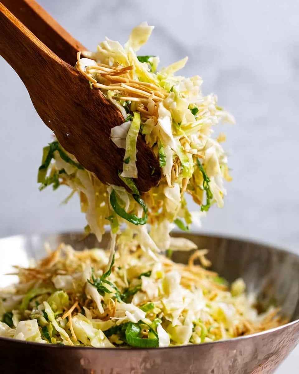 The image shows a fresh salad being lifted by two wooden spoons above a shiny metal bowl. The salad has three main layers: the bottom layer is pale green shredded lettuce with a soft, leafy texture; the middle layer consists of light brown crispy noodles adding crunch; and the top layer has thin green strips of herbs scattered throughout, giving a fresh touch. The background has a soft, white marbled texture, making the colors of the salad stand out clearly. photo taken with an iphone --ar 4:5 --v 7