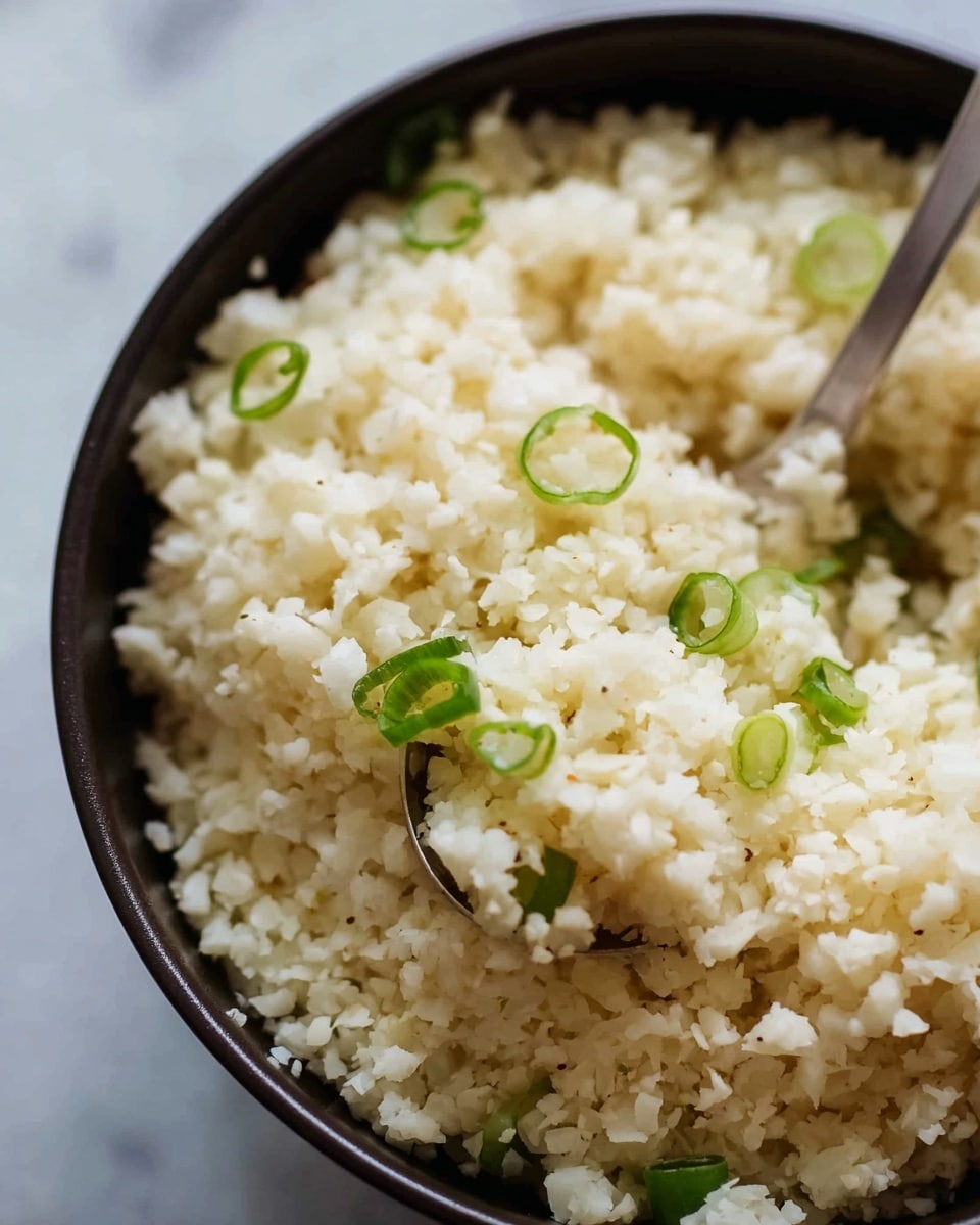 The image shows a close-up of a bowl filled with cooked rice, which is off-white and fluffy in texture. Scattered on top of the rice are thinly sliced green onion pieces, adding pops of fresh green color. A silver spoon with a rounded bowl is partially buried in the rice, ready to serve. The rice grains appear soft and slightly sticky, with some small clusters visible. The bowl holding the rice is white, and the whole scene rests on a white marbled texture surface. Photo taken with an iphone --ar 4:5 --v 7