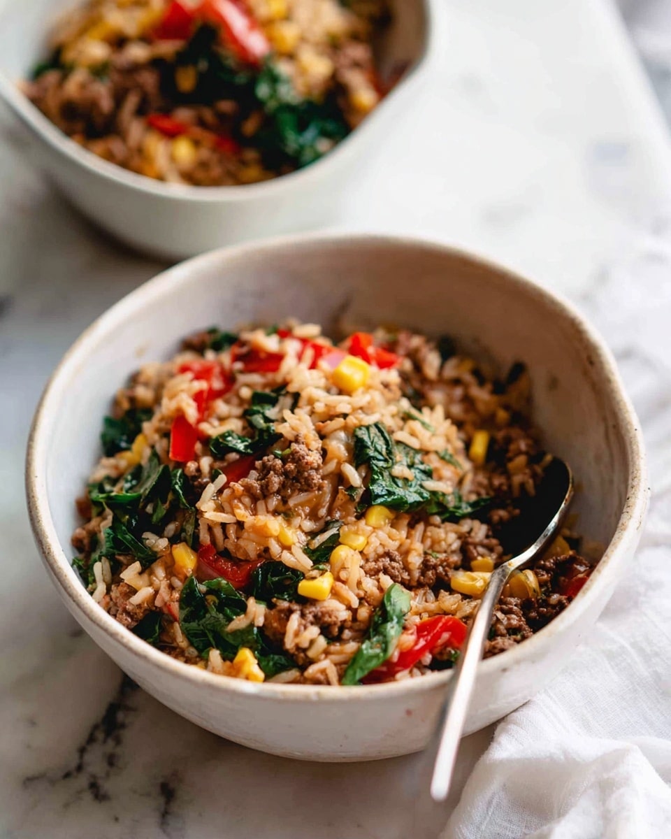 A close-up view of a white ceramic bowl filled with cooked rice mixed with minced meat, chopped red bell peppers, spinach leaves, and small bits of corn, all combined in a textured, savory blend that shows a mix of brown, red, green, and yellow colors. A silver spoon rests inside the bowl on the right side. In the soft-focused background, another white bowl containing the same dish is partly visible. The setup is on a white marbled surface with a white cloth nearby. Photo taken with an iphone --ar 4:5 --v 7