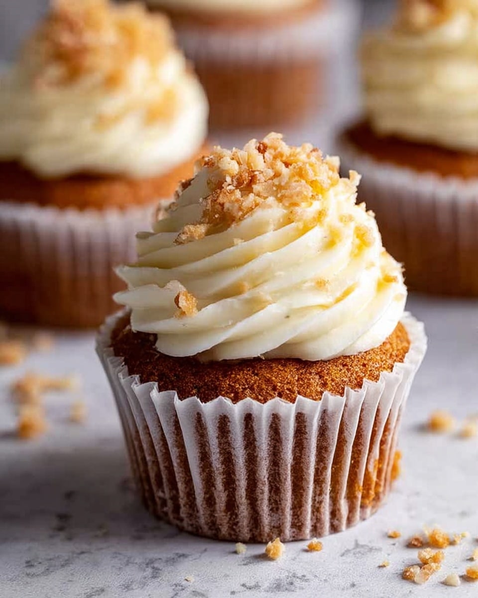 A close-up of a cupcake with three layers: the bottom is a dark brown cake wrapped in a white paper liner with visible creases, the middle layer is light cream-colored frosting swirled thickly in a spiral, and the top layer is sprinkled with small light brown nut pieces. The cupcake sits on a white marbled surface, with scattered nut crumbs around it. Two more similar cupcakes are blurred in the background. Photo taken with an iphone --ar 4:5 --v 7