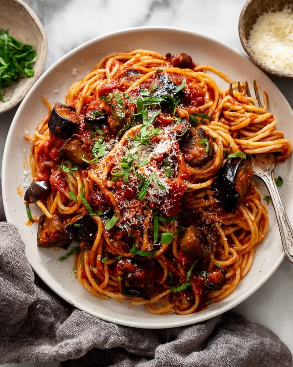 A white shallow bowl holds a serving of spaghetti pasta twisted into a small mound as the base layer, coated with bright red tomato sauce that drips down the noodles. On top, thick chunks of dark roasted eggplant are scattered, adding a deep purple color and chunky texture. The dish is sprinkled with grated white cheese and fresh green chopped herbs, adding contrast and freshness to the rich red and purple tones. A silver fork rests on the right side of the plate, partially under the pasta. The bowl sits on a soft gray cloth over a white marbled surface, with a small black dish of extra grated cheese nearby. Photo taken with an iphone --ar 4:5 --v 7