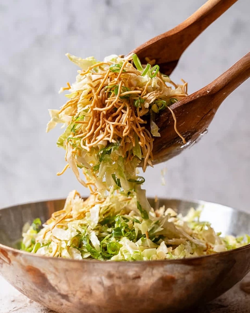 A close-up image shows wooden salad tongs lifting a fresh salad above a shiny metal bowl. The salad has several layers: shredded pale yellow cabbage forming the base, mixed with thin green leafy pieces and some crunchy light brown thin sticks on top. The wooden tongs hold a generous bite, with the salad looking fresh, crisp, and textured. The background is a white marbled surface with soft lighting. photo taken with an iphone --ar 4:5 --v 7