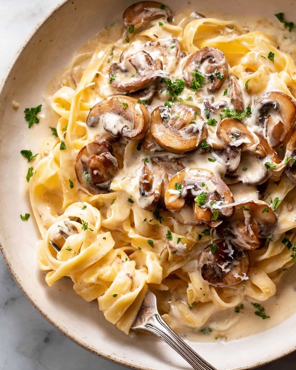 The image shows a close-up of creamy mushroom pasta on a white plate placed on a white marbled surface. The dish has three main layers: a base of thick, light yellow fettuccine noodles, followed by a generous mix of sliced brown mushrooms with a soft texture, all covered in a rich and creamy white sauce with a smooth and slightly glossy finish. The top is sprinkled with small green parsley pieces that add a fresh pop of color. At the bottom left, a silver fork is holding a bite of pasta and mushrooms, which also shows part of a woman's hand. Photo taken with an iphone --ar 4:5 --v 7