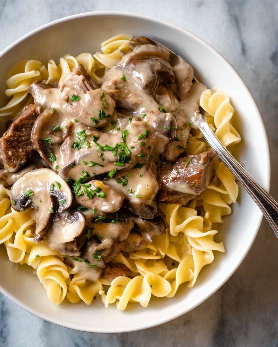 A close-up view of a white bowl filled with yellow spiral pasta at the bottom, topped with a creamy brown mushroom and beef sauce. The sauce covers the sliced mushrooms and pieces of beef, garnished with small green herbs sprinkled on top. A silver spoon rests inside the bowl on the right side, with the dish placed on a white marbled surface. photo taken with an iphone --ar 4:5 --v 7