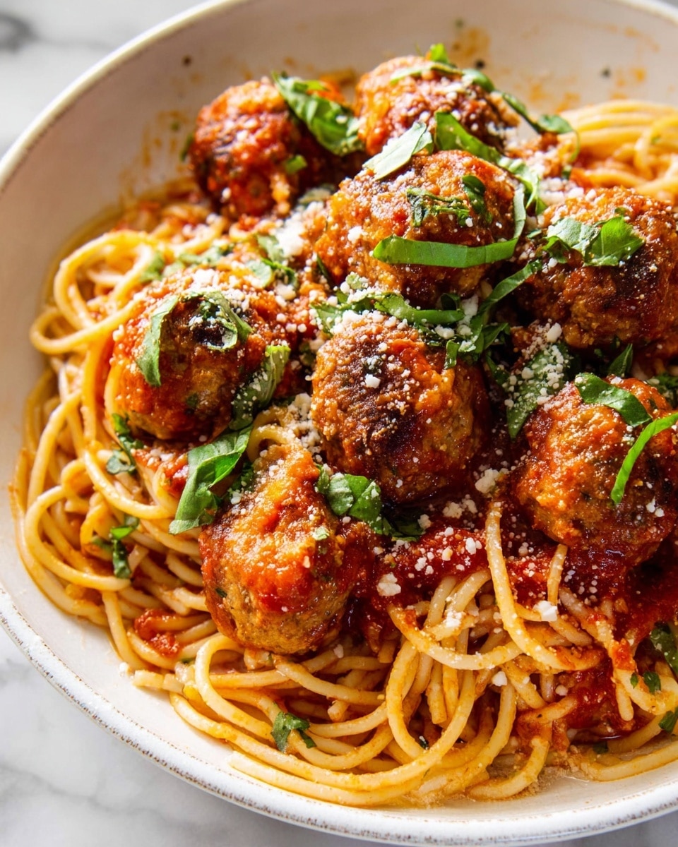 A close-up view of a white bowl filled with spaghetti and meatballs. The bottom layer contains long, light brown spaghetti noodles covered in bright red tomato sauce with bits of tomato visible. On top, there are several round, brown meatballs coated in sauce, sprinkled with small green basil leaves and white grated cheese. The texture of the noodles looks smooth and soft, while the meatballs have a crispy surface with some roughness. The white bowl has a simple design and sits on a white marbled surface. photo taken with an iphone --ar 4:5 --v 7