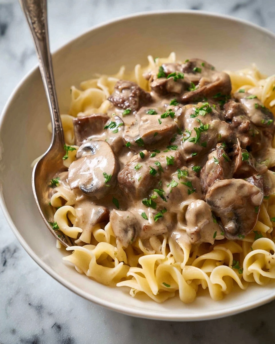A close-up view of a white bowl filled with a creamy meal of three main layers: at the bottom, there is twisted light yellow pasta neatly arranged; on top of that, there are slices of brown cooked mushrooms and pieces of browned meat; the top layer is covered with a light brown creamy sauce draped over the mushrooms and meat, speckled lightly with chopped green herbs, adding a touch of color. A silver spoon rests on the edge of the bowl. The bowl sits on a surface with a white marbled texture. photo taken with an iphone --ar 4:5 --v 7