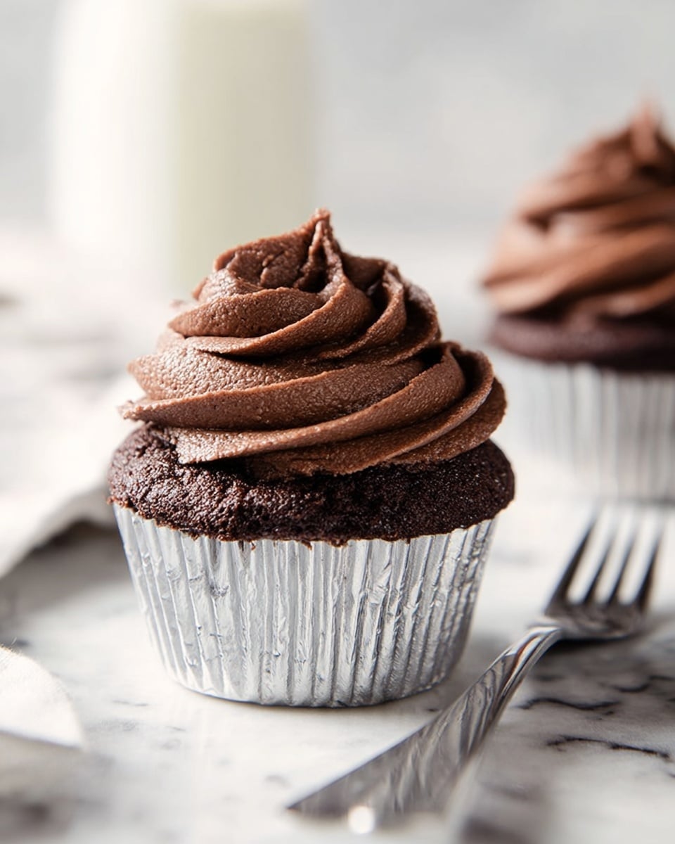 A close-up image of a single chocolate cupcake with two visible layers: the bottom layer is a dark brown, moist cake with a slightly crumbly texture, wrapped in a shiny silver foil liner, and the top layer is a generous swirl of rich, smooth, dark chocolate frosting with a light, fluffy texture, sitting neatly on top of the cake. The cupcake is placed on a white marbled surface, and in the blurred background, there are two metal forks and another cupcake with similar texture and frosting. The lighting is soft and natural, highlighting the creamy details of the frosting. Photo taken with an iphone --ar 4:5 --v 7