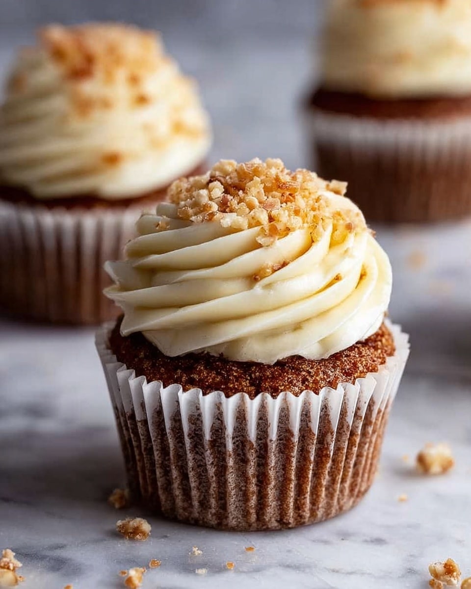 A close-up of a single cupcake with a light brown base wrapped in a white paper liner showing a slightly rough texture. On top is a thick swirl of creamy white frosting with a smooth, soft texture, piled in about three layers. The frosting is sprinkled with small chopped light brown nuts, some scattered on the white marbled surface around the cupcake. Two more cupcakes with similar colors and frosting are blurred in the background. photo taken with an iphone --ar 4:5 --v 7
