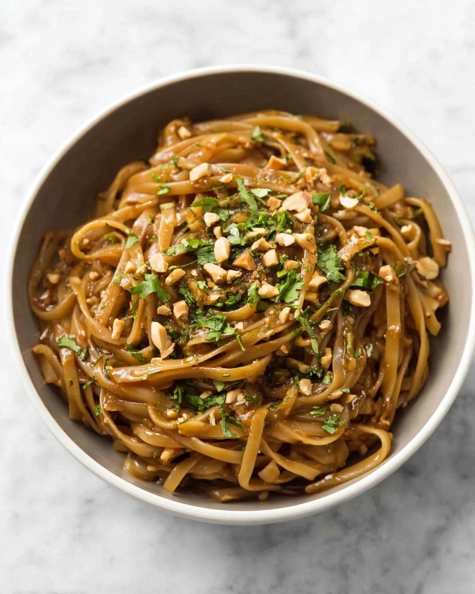 A dark bowl holds a serving of wide, flat noodles coated in a thick, light brown sauce. The noodles are mixed with small bits of vegetables and sprinkled with crushed peanuts and finely chopped green herbs. The noodles are piled in a loose mound with the sauce clinging to their shiny surface. The photo is set on a white marbled surface. photo taken with an iphone --ar 4:5 --v 7